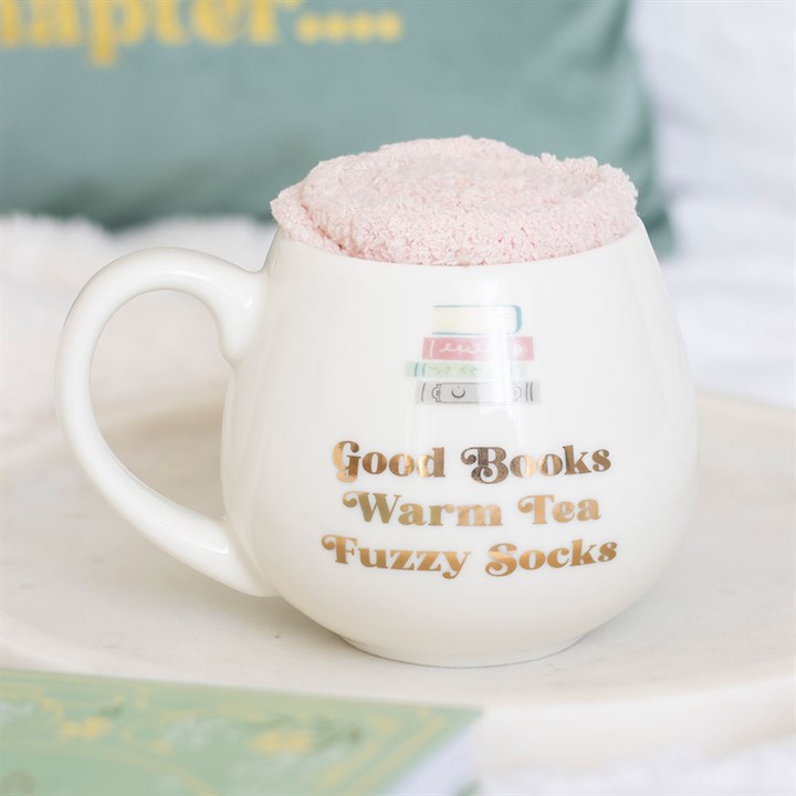 White mug with text 'Good Books Warm Tea Fuzzy Socks' and a pink fluffy object on top, on a soft surface.