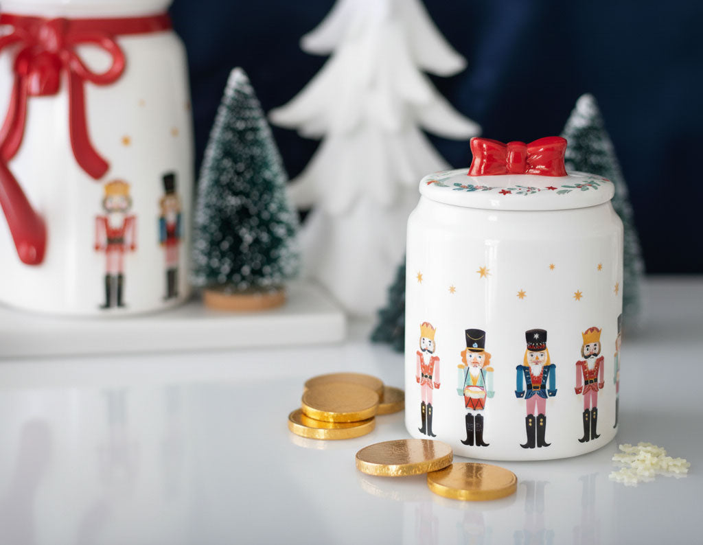Decorative cookie jar with nutcracker design on a table with gold coins and Christmas trees in the background.